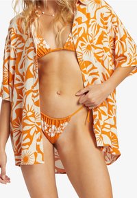Koszula w kwiaty w odcieniach pomarańczowego i kremowego noszona na pomarańczowym bikini; koszula ma krótkie rękawy oraz luźny krój, z zapięciem na guziki i dekoltem w kształcie łódki.