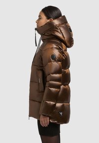 Brūna pūkaina jaka ar spīdīgu, šūtu tekstūru, pārmērīgi lielu kapuci, sānu kabatām un melnu logotipa uzlīmi uz piedurknes. Izstrādāta siltumam.