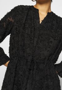 Vestido negro con tejido texturizado y esponjoso, con escote en V, mangas largas y una cintura con cordón para un ajuste ajustable.