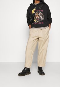 Person som bär beige byxor, svarta kängor, svart hoodie med grafisk av en rytande tiger och en svart crossbody-väska, står med händerna i fickorna.