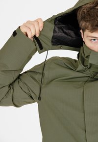 Olivgrüne Jacke mit hohem Kragen, verstellbarem Kordelzug und schwarzem Innenfutter. Strukturiert mit sichtbaren Nähten und Klettverschluss.