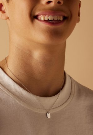 Zilveren ketting met een rechthoekige tag hanger die op een lichtgrijs shirt rust, met nadruk op het gladde metaaloppervlak en het eenvoudige ontwerp.