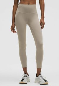 Beige high-waisted leggings van glad, rekbaar materiaal met platte naden, ontworpen voor een comfortabele pasvorm, gecombineerd met wit en zwarte sportschoenen.