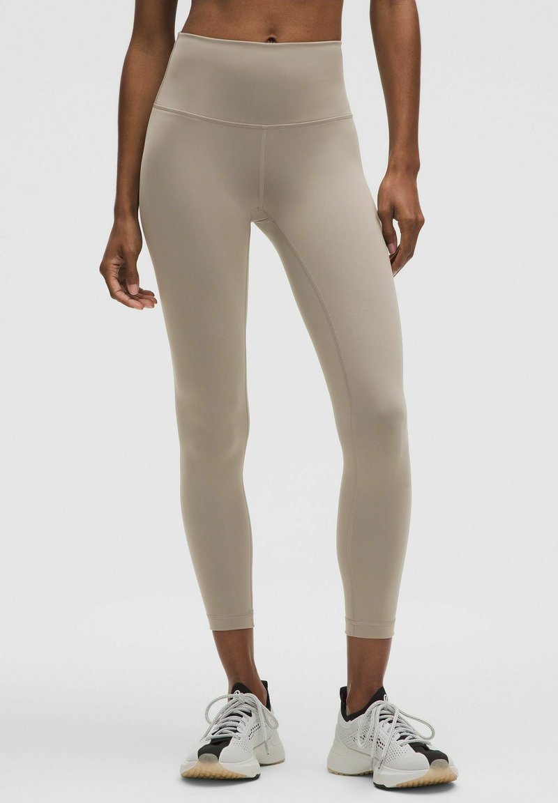Beige high-waisted leggings van glad, rekbaar materiaal met platte naden, ontworpen voor een comfortabele pasvorm, gecombineerd met wit en zwarte sportschoenen.