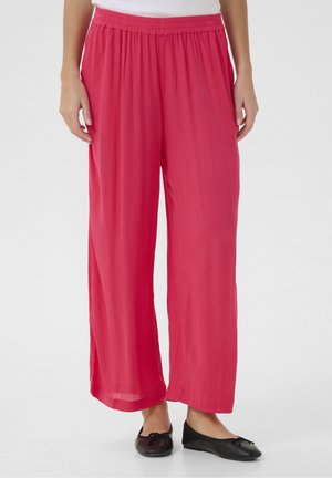 Frau trägt lockere, knallpinke Hosen mit elastischem Bund und schwarze Ballerinas, steht vor einem schlichten weißen Hintergrund.