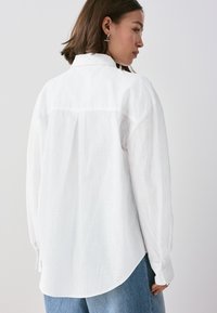 Weißes Langarmhemd mit lockerem Schnitt, dezentem Strukturmuster, abgerundetem Saum und geknöpften Manschetten. Kombiniert mit hellblauen Jeans.