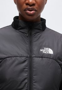Gros plan d’une personne portant une veste noire North Face avec une fermeture éclair à l’avant et un logo visible sur le côté gauche de la poitrine.
