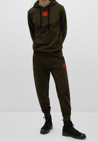 Olivgrünes Hoodie- und Jogginganzugs-Set mit einer vorderen Kängurutasche und rotem Logoaufnäher. Kombiniert mit schwarzen, strukturierten Stiefeletten.