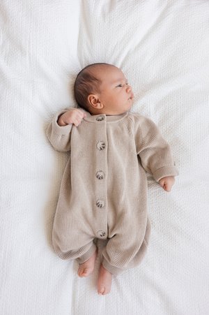 Romper beige in maglia con maniche lunghe e cinque bottoni, indossato da un bebè sdraiato su una coperta bianca testurizzata. Materiale morbido e vestibilità rilassata.