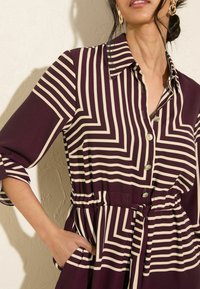 Bordeauxfarbige Bluse mit weißen Zickzackstreifen, Knopfleiste, Hemdkragen, elastischem Bund und seitlichen Taschen. Weicher Stoff mit glatter Textur.