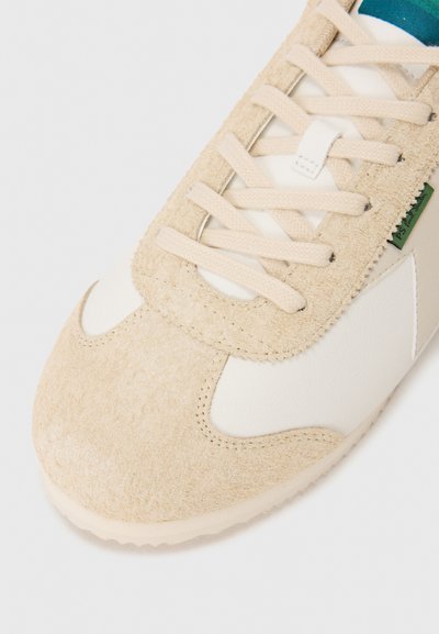 Sneaker med en beige semsket overdel og hvite lærdetaljer, teksturerte snørebånd og en glatt gummisåle. Minimalistisk design med grønn logo.