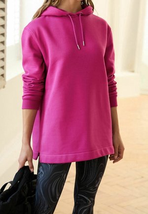 Personne portant un sweat à capuche rose vif avec cordons et des leggings noirs à motifs, tenant un sac à dos noir dans un espace intérieur lumineux.