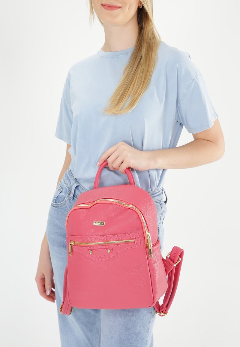 Sac à dos en simili cuir rose avec des zippers dorés, de forme arrondie, poche avant et anse supérieure. Porté par une personne en chemise bleu clair.