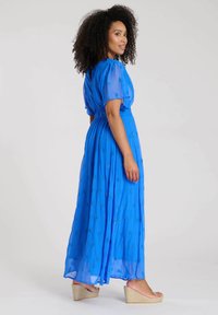 Lång blå chiffongklänning med korta puffärmar, blommönster, elastisk midja och flödande silhuett. Bärs med kilklackssandaler.