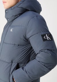Muž oblečen�ý v podšité modré bundě Calvin Klein s kapucí, logy na hrudi a rukávu, ruka v kapse, mírně otočený vlevo.