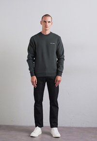 Man stands against plain wall wearing dark green sweatshirt, black pants, and white sneakers, facing forward with arms relaxed.