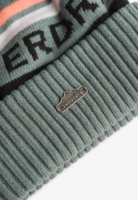 Dzianinowa czapka w jasnym zielonym kolorze z prążkowanym wzorem. Posiada czarno-koralowy paskowany wzór oraz srebrną metkę z logo Superdry.