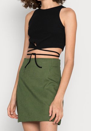 Femme portant un haut court noir sans manches avec des liens à nouer autour de la taille et une mini-jupe à motif pied-de-poule verte, sur un fond uni.