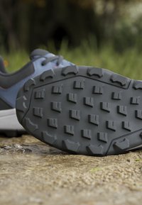 Semelle d'une chaussure de randonnée bleue et grise avec des crampons profonds marchant sur un sol humide et rocheux dans un cadre naturel en plein air.