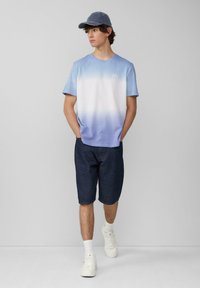 Helltürkis zu weißem Farbverlauf T-Shirt, kombiniert mit dunklen Denim-Shorts. Weiße Sneaker und eine Jeansmütze runden das Outfit ab. Einfaches Design.