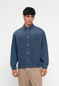 Denim jakna v temno modri barvi, z gumbnim zapiranjem in ovratnikom, ena prsna žepta in dolgi rokavi. Ima kovinske gumbe in sproščen kroj.