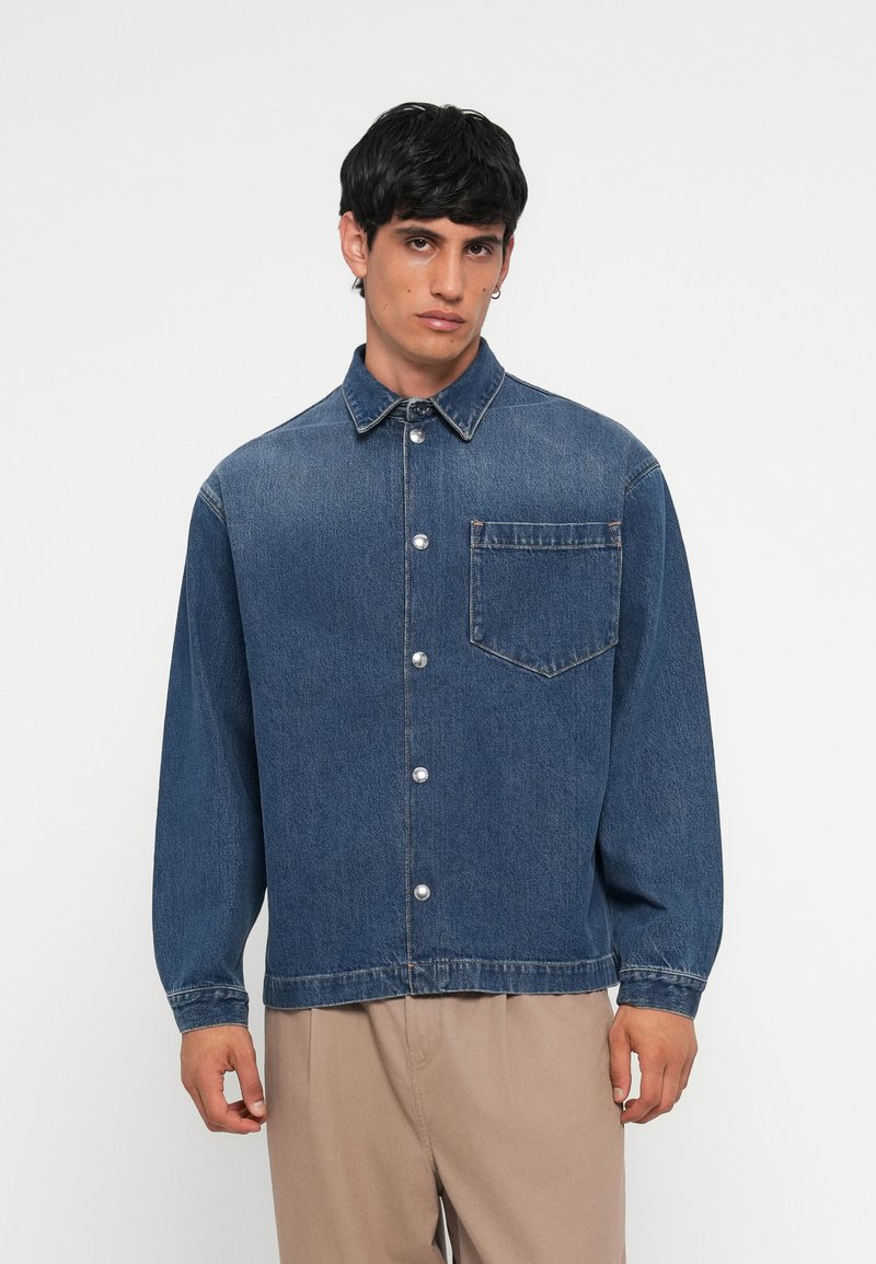 Denim jakna v temno modri barvi, z gumbnim zapiranjem in ovratnikom, ena prsna žepta in dolgi rokavi. Ima kovinske gumbe in sproščen kroj.