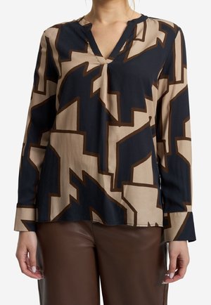 Vrouw draagt een blouse met lange mouwen en een V-hals, voorzien van een gedurfd abstract marineblauw en beige geometrisch patroon, en bruine leren broek.
