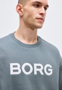 Άνδρας που φοράει γκρι φούτερ με μεγάλο λευκό κείμενο "BORG", με μικρό κρίκο στο αριστερό αυτί, κοιτάζει δεξιά σε απλό φωτεινό φόντο.