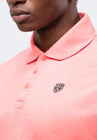 Κοντινό πλάνο ενός ατόμου που φοράει μοβ ροζ πόλο με μικρό μαύρο-άσπρο κεντητό λογότυπο στο στήθος.