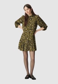 Blumenkleid in Olivgrün und Beige mit langen Puffärmeln, einem Rüschensaum und einem gerafften Ausschnitt. Kombiniert mit schwarzen Schuhen.