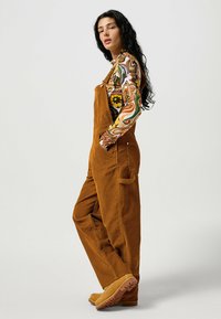 Brown corduroy overalls with a relaxed fit, featuring wide legs, side pockets, and adjustable straps. Paired with a colorful patterned long sleeve shirt.