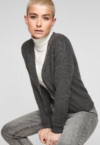 Cardigan en tricot gris avec un décolleté en V profond et des manches longues, porté sur un col roulé blanc. Présente un tissu texturé et des accents de boutons.