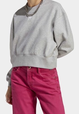 Person wearing a grey cropped sweatshirt with a silver chain necklace and high-waisted bright pink pants, standing with one hand visible.