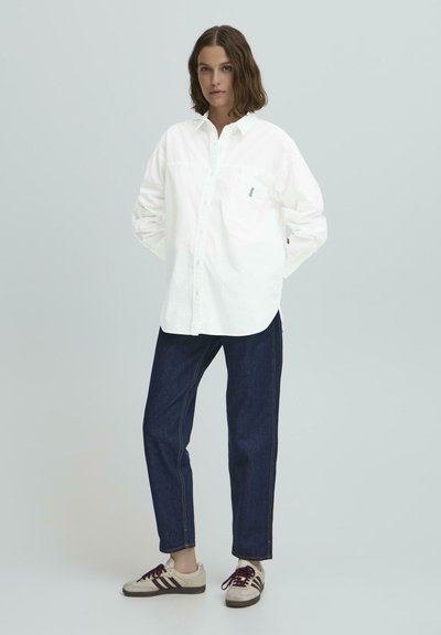 Balta marškiniai su ilgomis rankovėmis ir krūtinės kišene, deranti su tamsiai mėlynomis džinsomis ir kreminiais sportbačiais su bordo juostelėmis.