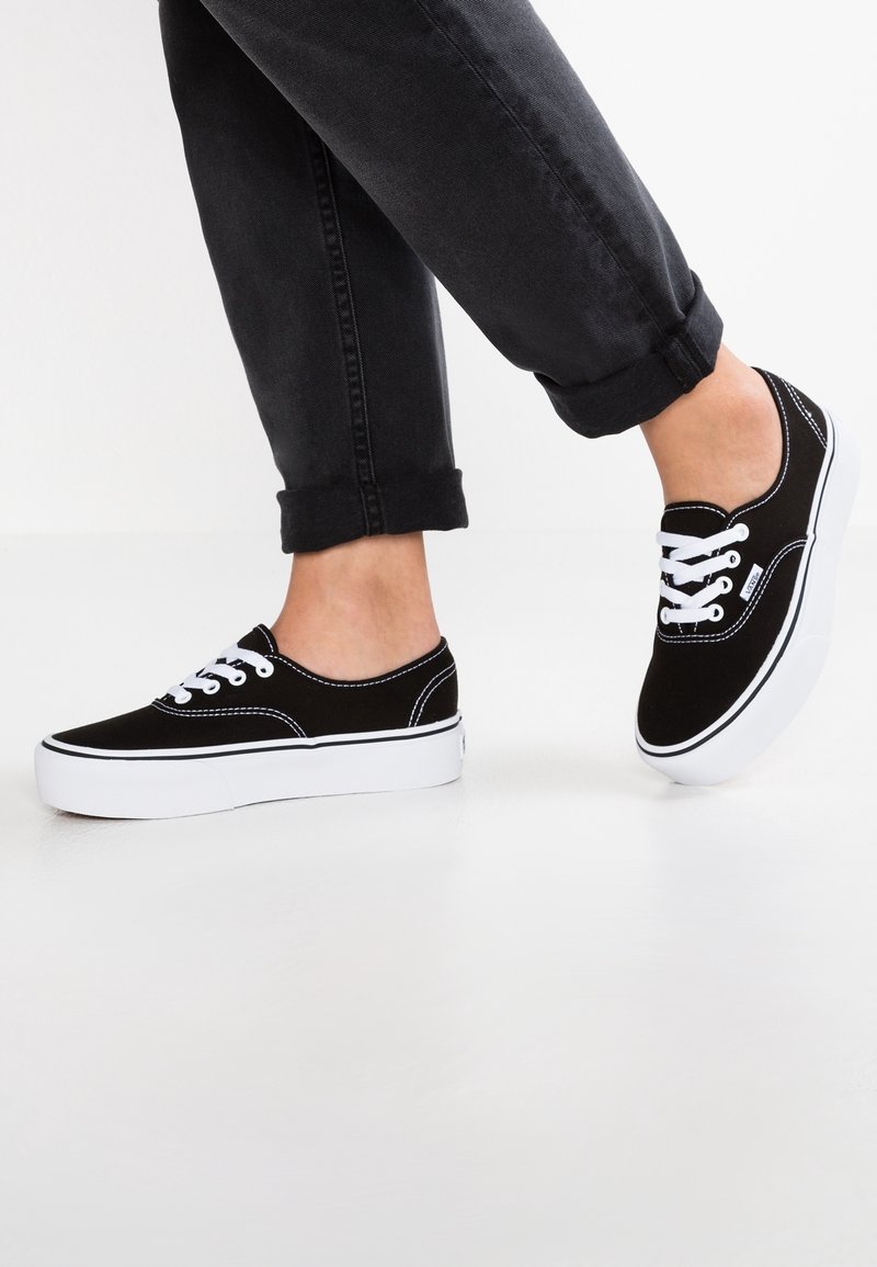 Personne portant des baskets en toile noires avec des lacets et semelles blanches, debout sur un sol blanc avec un pantalon noir retroussé.