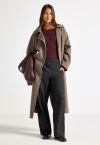 Kvindelig iført en lang brun frakke, burgunder top, mørke ternede bukser, holder en stor burgunder tote bag, står foran en ensfarvet hvid baggrund.