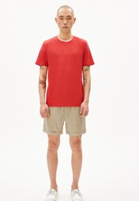 Rode katoenen t-shirt met ronde hals, gedragen met beige linnen shorts met opgerolde mouwen. Model staat met de handen naast zich.