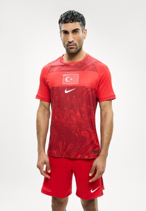 Άνδρας που στέκεται φορώντας κόκκινη φανέλα και σορτς ποδοσφαίρου της Τουρκίας με λογότυπα Nike σε απλό λευκό φόντο.