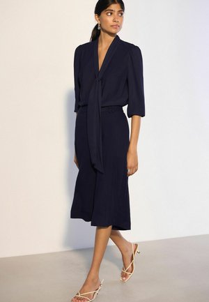 Frau trägt ein dunkelblaues Midikleid mit Dreiviertelärmeln und einer vorderen Schlinge, kombiniert mit beigen Riemchen-Pumps mit Absatz, steht drinnen.