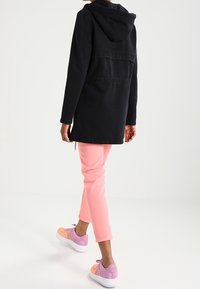 Mustat hupullinen takki, joka on pitkä, yhdistettynä vaaleanpunaisiin kapeneviin housuihin ja monivärisiin lenkkareihin, joissa on violetteja ja persikan sävyjä.