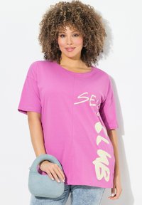 Una camiseta de manga corta rosa con cuello redondo y texto gráfico en color crema. Un bolso azul claro con un asa complementa el conjunto.