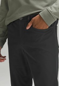Crne hlače od glatkog, rastezljivog materijala slim fit kroja, s bočnom džepom i kopčanjem na gumbe na struku.
