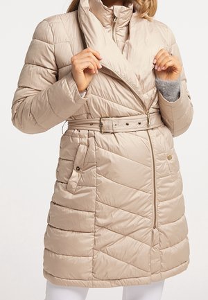 Mujer con abrigo acolchado beige con cinturón, costuras diagonales y cremallera frontal, ajustándose el cuello.