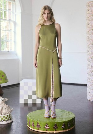 Oliivivihreä hihaton mekko, jossa on kukkareunus, polvipituinen halkio, yhdistettynä keltaisiin nauhakorkokenkiin ja vaaleanpuniin sukkiin, pyöreällä vihreällä alustalla.