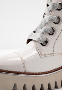 Valkoinen kiiltonahkainen saapas, jossa paksu platakorko, harmaat nupukkinauhat, metalliset kengännauhareiät ja kontrastiväriset ompeleet.