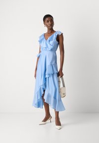 Light blue dress featuring ruffled sleeves, a V-neckline, and tiered hem. Model holds a small cream handbag, wearing white high-heeled shoes.
