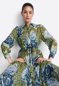 Šaty s kvetinovou textúrou v modro-zelených odtieňoch, s golierovým výstrihom, zapínaním na gombíky vpredu, elastickými rukávmi a sťahovaným pásom so zaväzovaním.