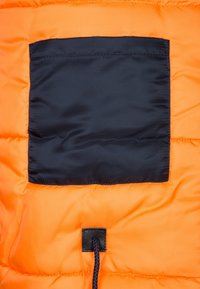 Interno di una giacca a vento arancione con una tasca quadrata nera e un cordino nero con un togglè nella parte inferiore. Tessuto testurizzato e isolato.