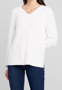 Pull blanc en maille avec col en V, manches longues, texture côtelée et fentes latérales. Associé à un jean bleu foncé.