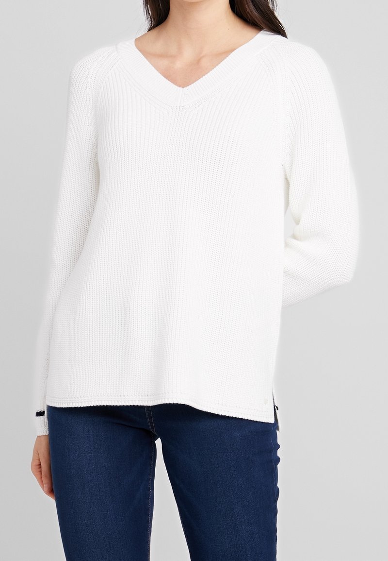 Pull blanc en maille avec col en V, manches longues, texture côtelée et fentes latérales. Associé à un jean bleu foncé.